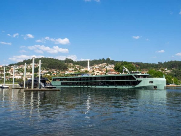 A sleek, modern green vessel for a luxury Nile River cruise docked in a peaceful river setting with lush green hills in the background