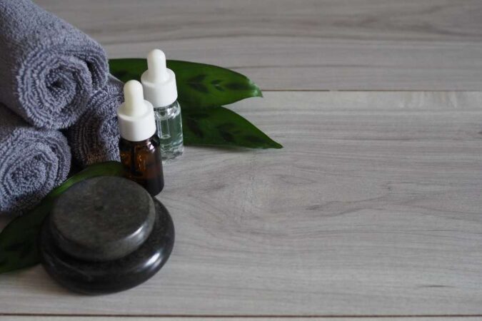 Stacked black basalt stones, dropper bottles of oil, and folded grey towels arranged on a wooden surface for a hot stone massage.